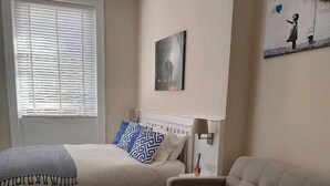 Een slaapkamer met een bed, een bank, een raam met zonwering, een schilderij aan de muur en een lamp op het nachtkastje.