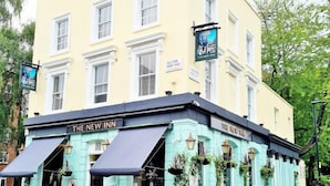 Een hoekpand met een blauwgroene gevel, genaamd 'The New Inn'. Het heeft een zwart dakzeil boven de ingang, terras met tafels en stoelen, en een bord met de tekst 'The Merry Inn' boven de ingang.