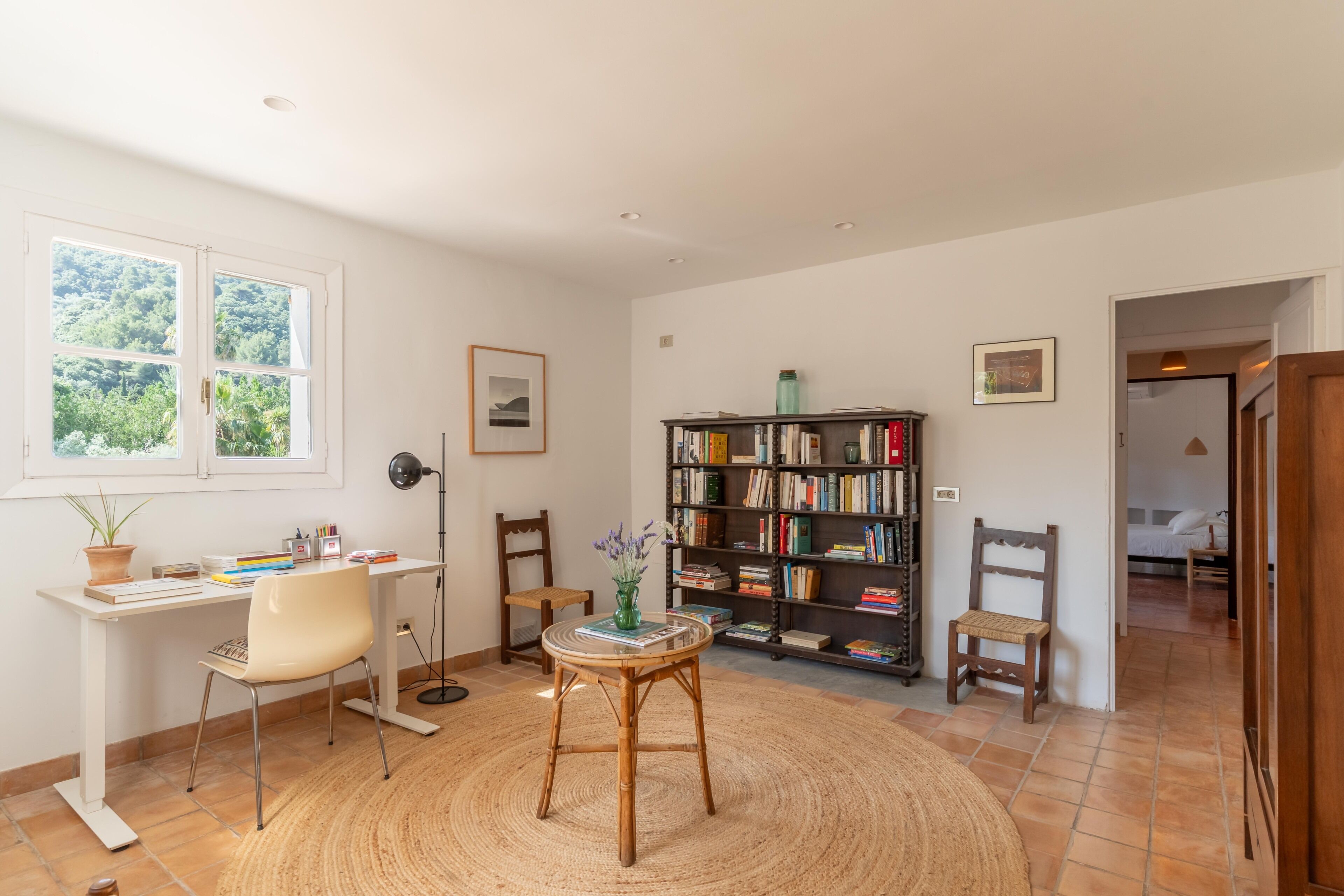 A room with a desk, chairs, a bookshelf, and a window with a view of greenery.