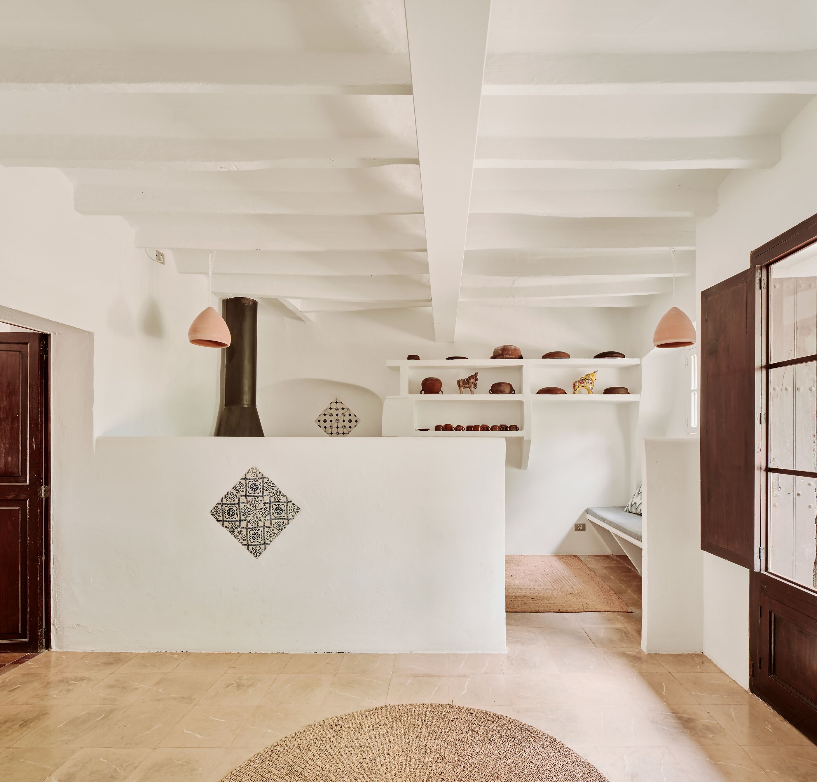 A room with a white wall, a wooden door, a patterned wall decoration, a wooden floor, a woven rug, and a shelf with pottery.