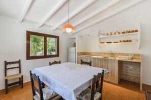 Uma sala de jantar com mesa, cadeiras, luminária pendente, janela e área de cozinha com prateleiras.