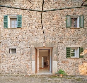 Um edifício de pedra com uma porta, duas janelas com portões e um pequeno espaço de entrada com plantas em vasos.