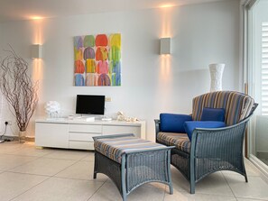 A modern living room with a flat-screen TV, a colorful abstract painting, a wicker chair with a striped cushion, and a small side table.