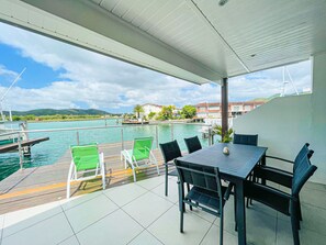A covered outdoor area with a table and chairs, overlooking a body of water with boats and buildings in the background. - Beautiful Waterfront Villa (Jolly Harbour)