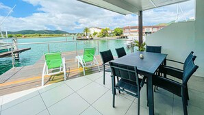 Un espace extérieur abrité, doté d’une table et de chaises, qui offre une vue sur une étendue d’eau où l’on aperçoit des bateaux et des bâtiments au loin.