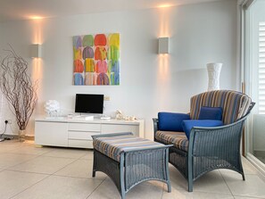 Una sala de estar moderna con un televisor de pantalla plana, un cuadro abstracto colorido, una silla de mimbre con un cojín a rayas y una pequeña mesita auxiliar.