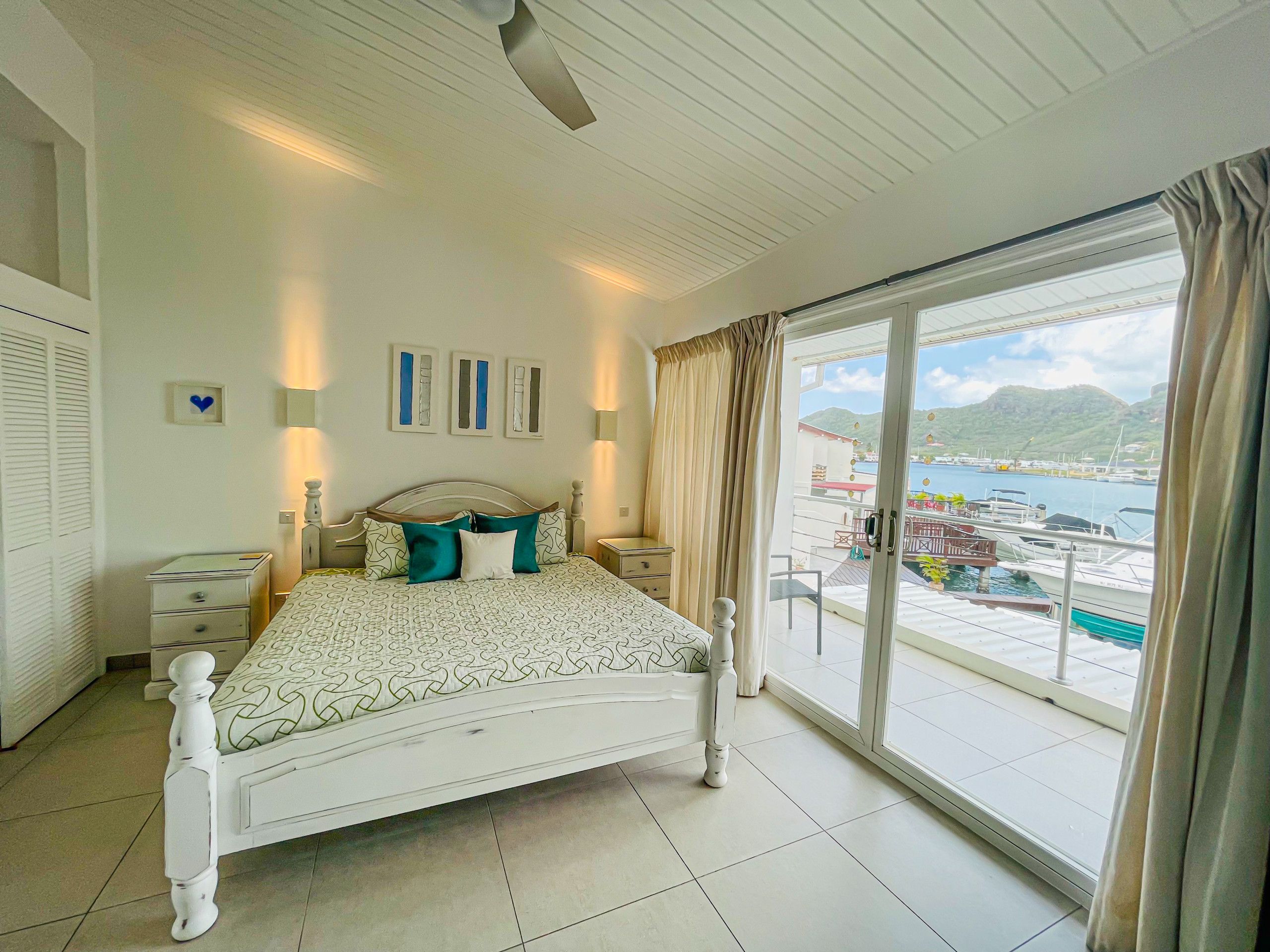 Ein Schlafzimmer mit Bett, Nachttischen, Kommode und großem Fenster mit Blick auf einen Yachthafen und Boote.