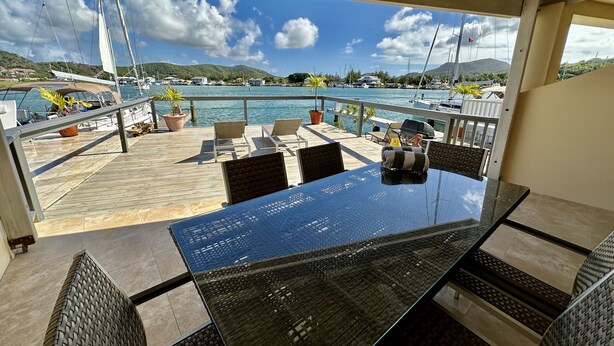 A covered outdoor seating area with a glass table and chairs, overlooking a marina with boats and a mountainous landscape. - Modern Waterfront Villa (Jolly Harbour)