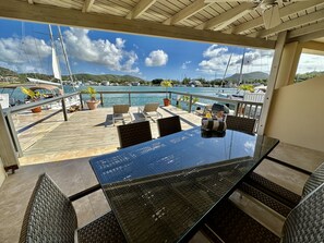 A covered outdoor seating area with a glass table and chairs, overlooking a marina with boats and a mountainous landscape. - Modern Waterfront Villa (Jolly Harbour)