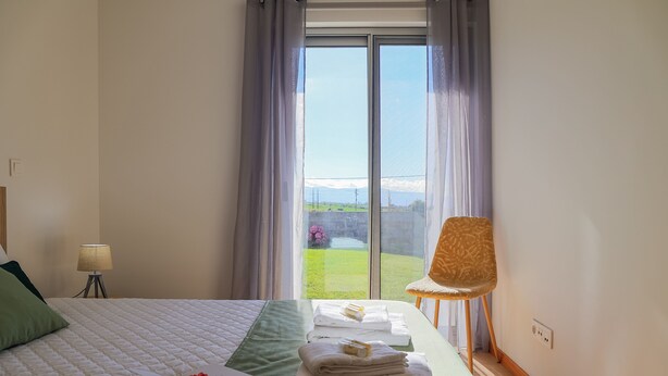 A hotel room with a large bed, a chair, a lamp, and a view of a green landscape through a window.
