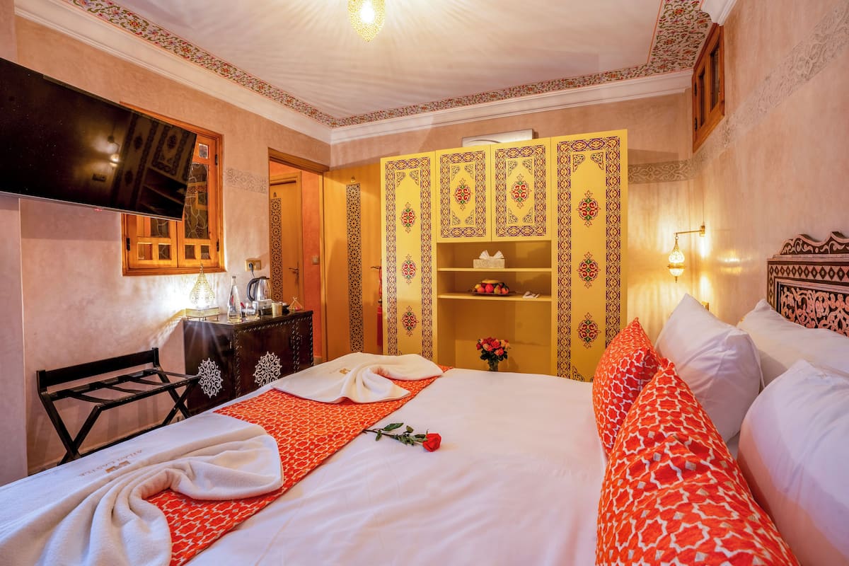 A neatly made bed with a red and white patterned bedspread, a rose, and a towel.