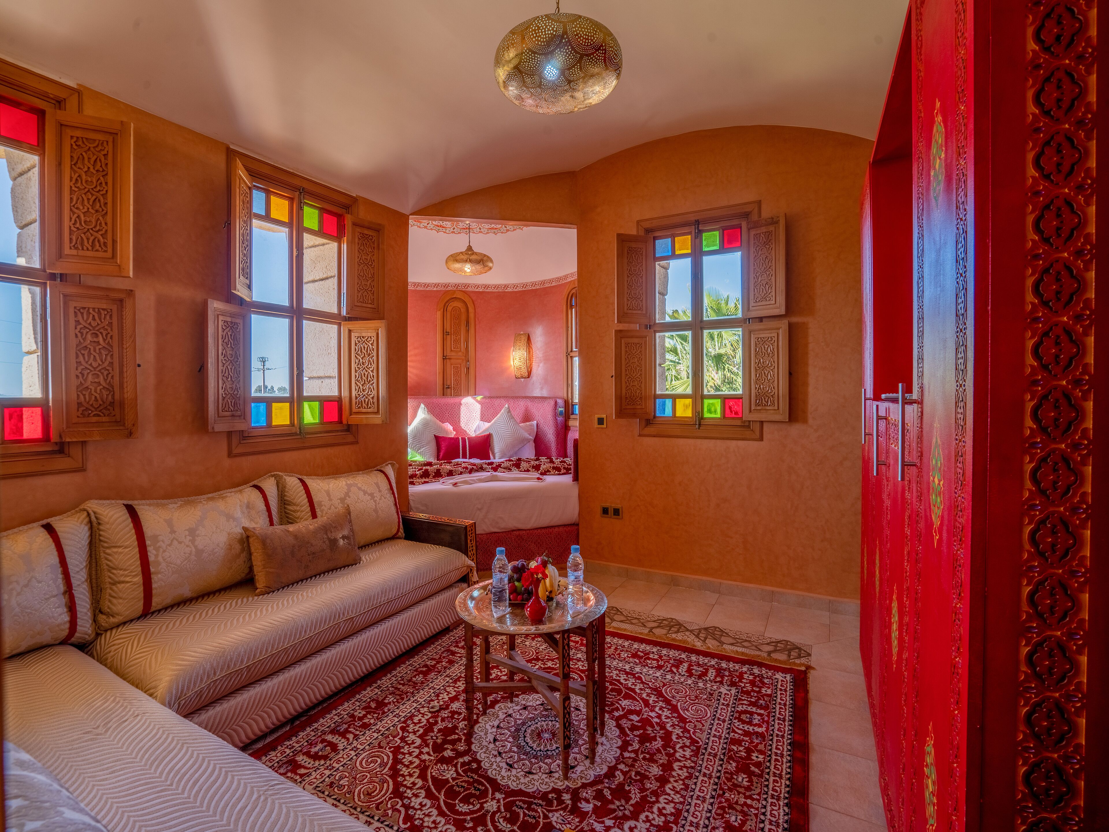 A room with a sofa, a round table with a red and white patterned rug, and a bed with a red headboard.