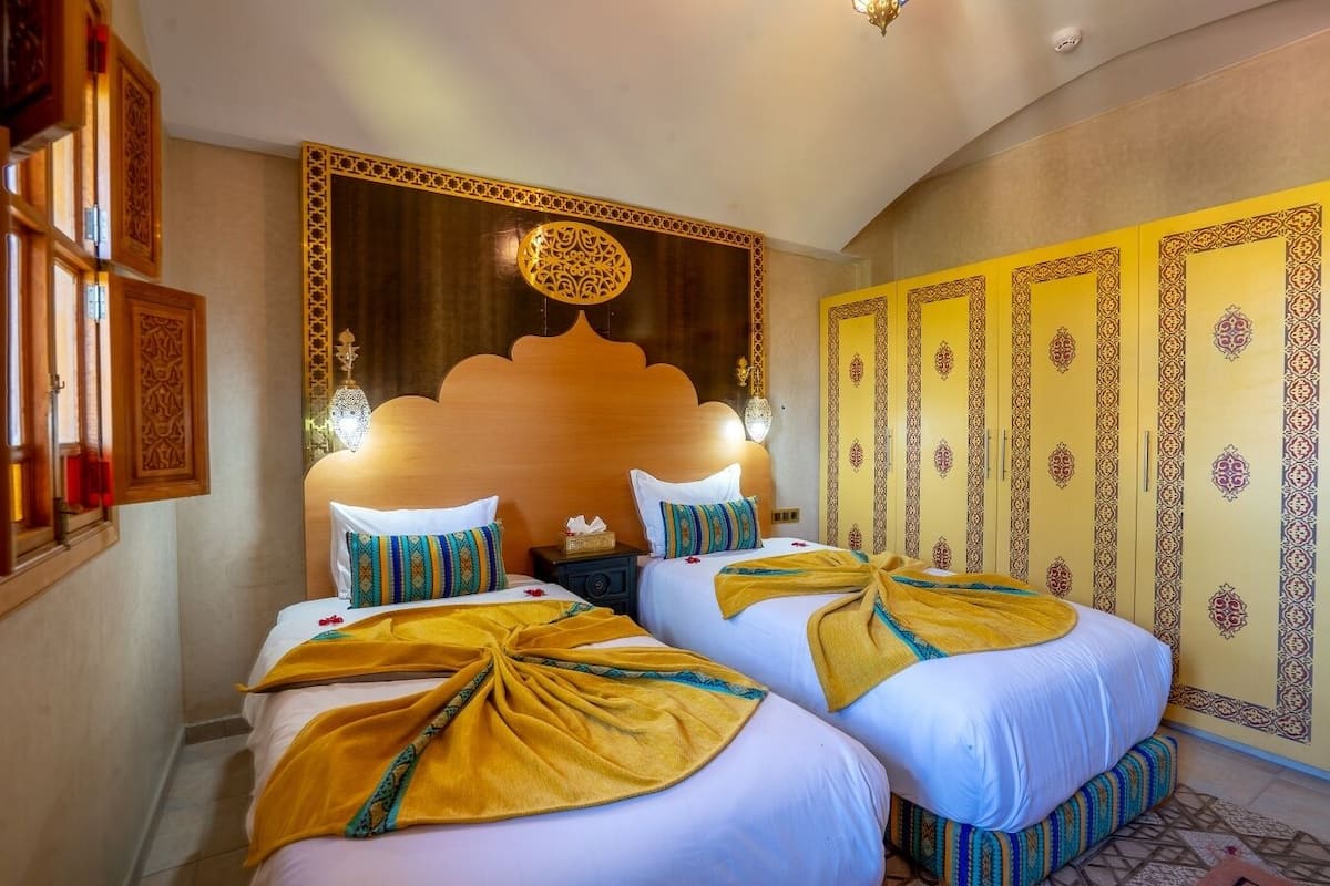 A hotel room with two beds, a decorative headboard, a wall with patterned panels, and a chandelier.