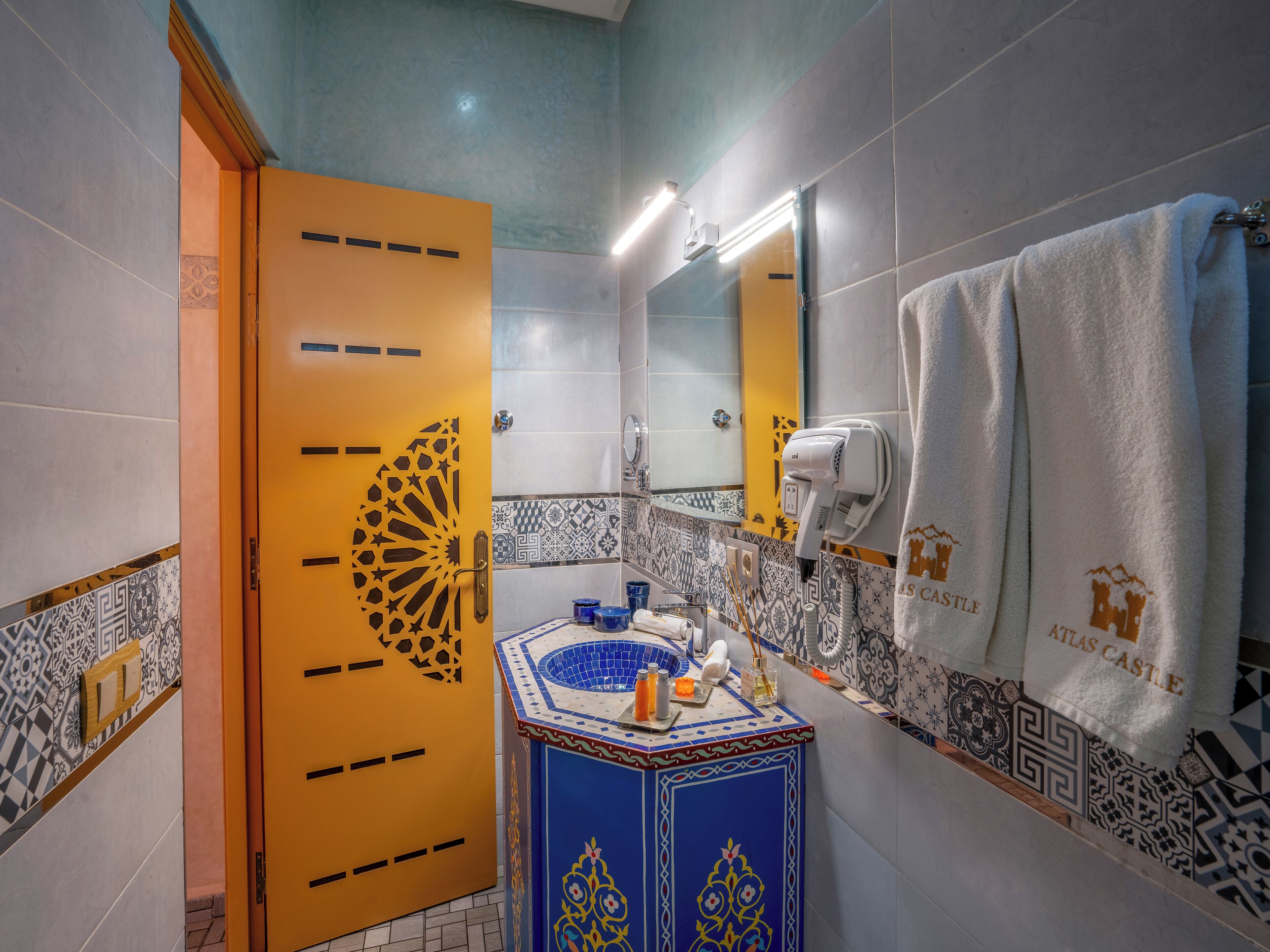 A bathroom with a yellow door, a sink with a blue and gold design, a towel rack with a white towel, and a hairdryer.