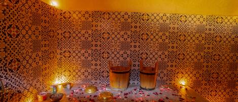 A room with patterned tiles, wooden barrels, candles, and scattered rose petals.