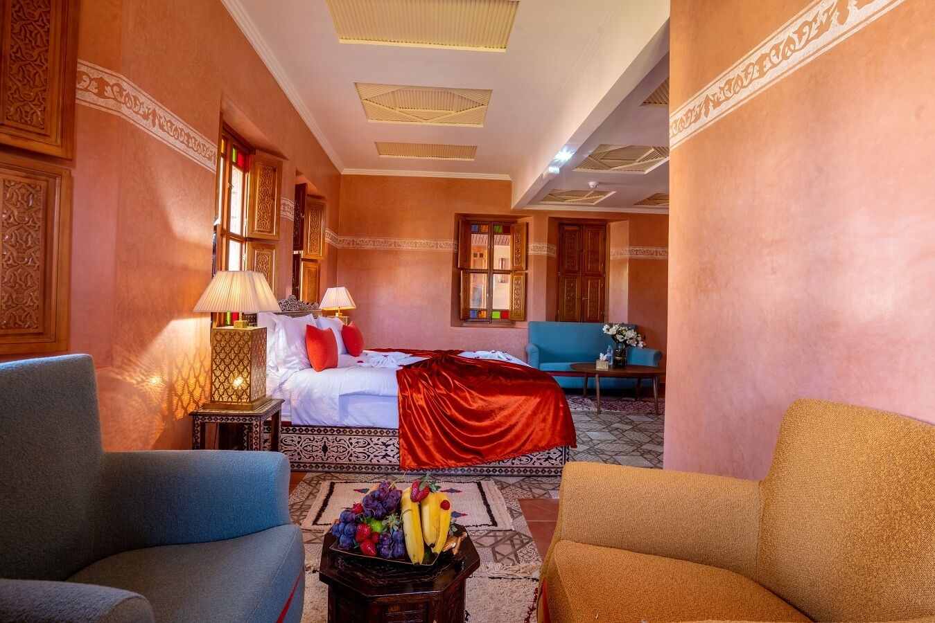 A hotel room with a bed, two chairs, a table with fruit, and a blue sofa.
