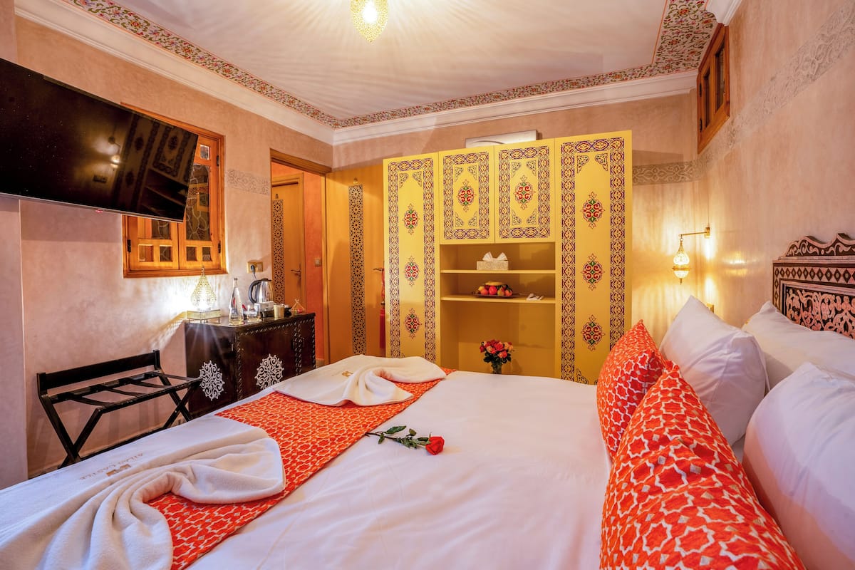 A neatly made bed with a red and white patterned bedspread, a rose, and a towel.