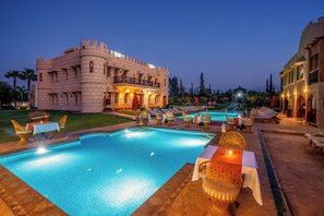 A swimming pool with lounge chairs and tables, a castle-like building, and a well-lit patio area.