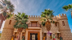 A castle-like building with a central entrance, palm trees, and mannequins dressed in traditional attire. - Atlas Castle Marrakech (Ghmate)