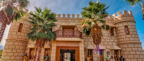 A castle-like building with a central entrance, palm trees, and mannequins dressed in traditional attire.