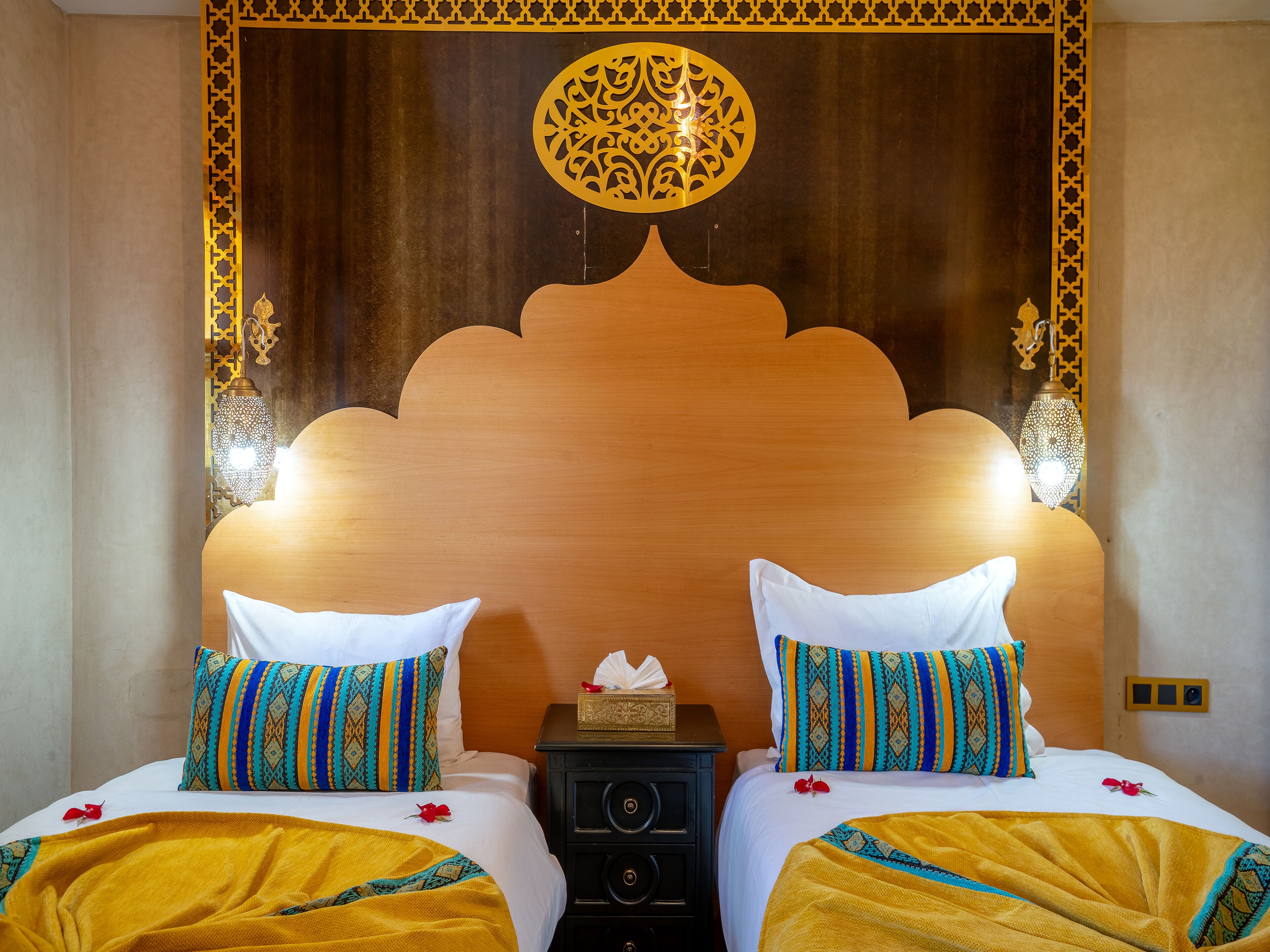Habitación de hotel con dos camas, cada una con almohadas estampadas y una manta amarilla. Hay una mesita de noche con una caja de pañuelos y una lámpara decorativa.