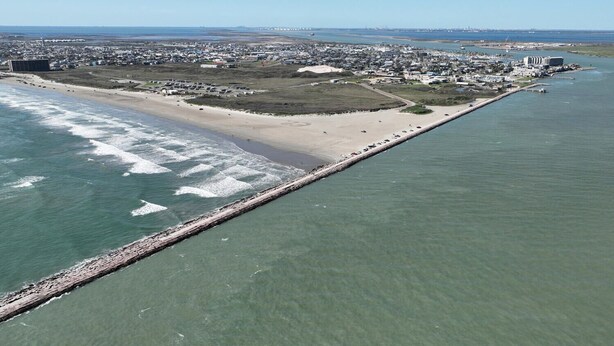 An aerial view of a coastal city with a long pier extending into the water, a sandy beach, and a densely populated urban area in the background.