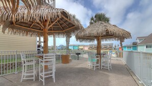 A covered outdoor seating area with white chairs and tables, surrounded by a white fence, under thatched umbrellas, and palm trees in the background.