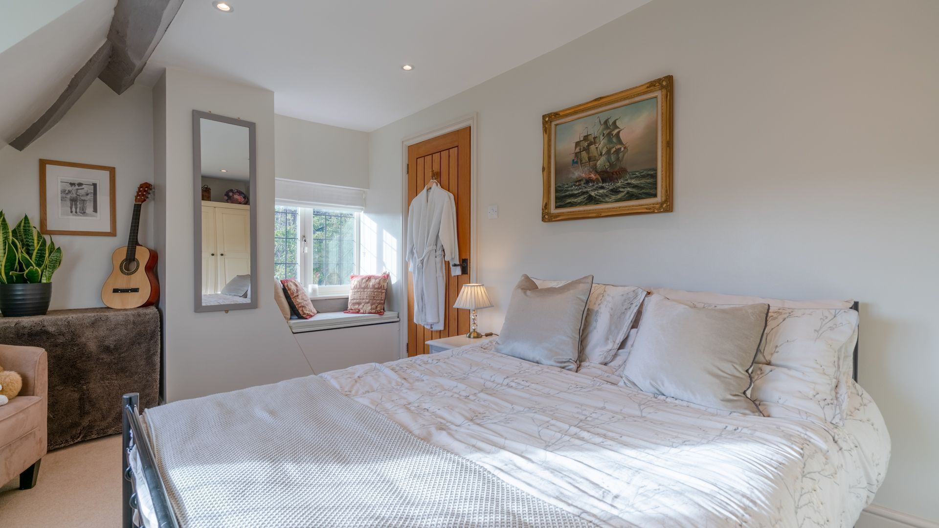 Een slaapkamer met een groot bed, een raam met uitzicht, een gitaar, een spiegel en een schilderij aan de muur.