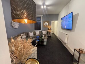 A modern living room with a sectional sofa, a coffee table, a TV mounted on the wall, and a decorative lamp. - Urban 3 Bedroom Home in Kings Heath-Great Location (King's Heath)