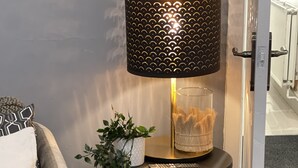 Een hoek van een kamer met een grijze bank, een klein rond tafeltje met een lamp, een potplant en een boekenkast vol boeken.