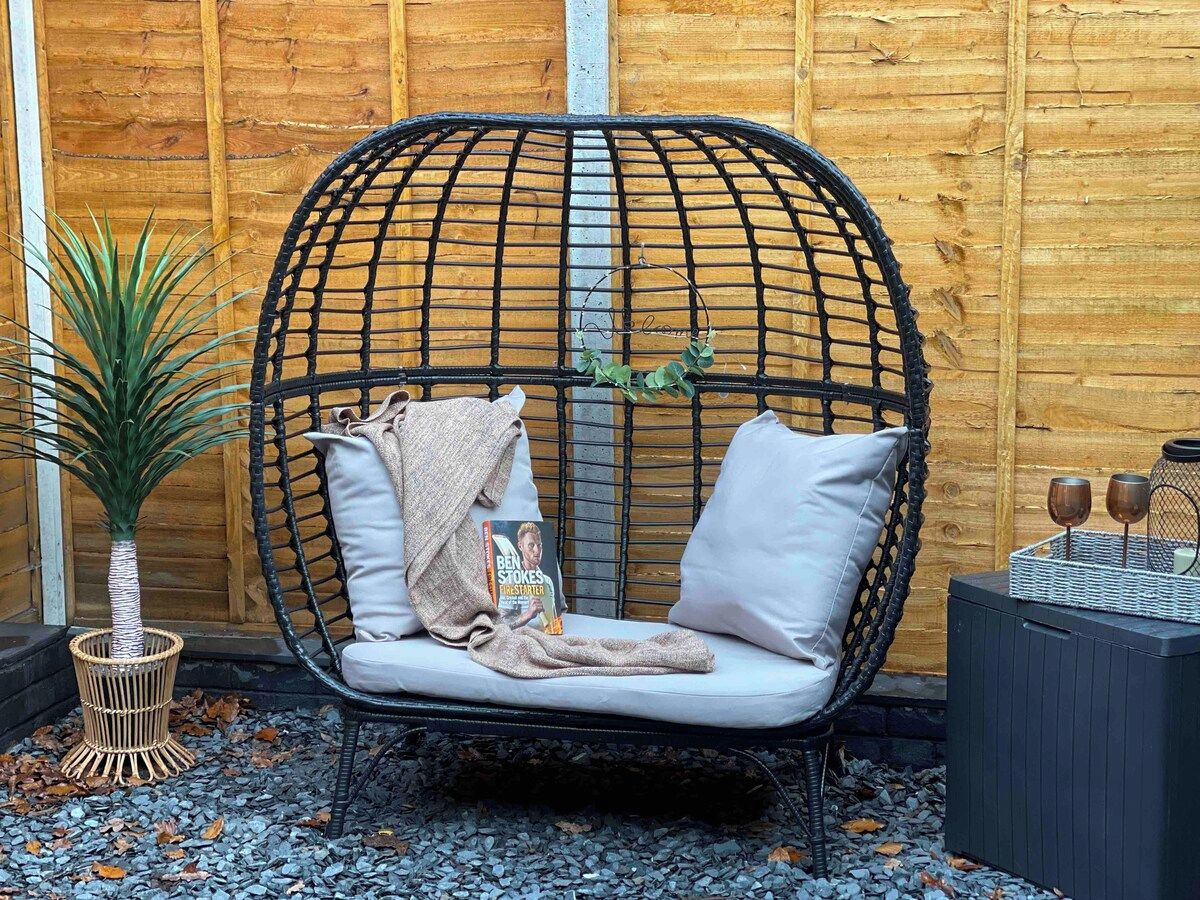 Une chaise en osier avec des coussins et un livre, posée sur une surface recouverte de petits cailloux, avec une plante à côté.