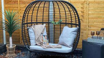 Une chaise en osier avec des coussins et un livre, posée sur une surface recouverte de petits cailloux, avec une plante à côté.