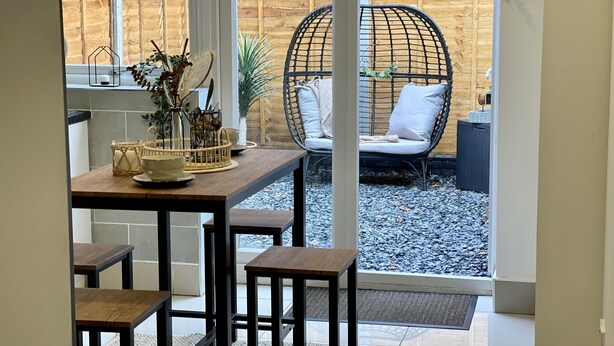 Een eethoek met een houten tafel en krukken, een rieten stoel met kussens, en zicht op een houten hek en een grindpad buiten.