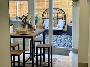 A dining area with a wooden table and stools, a wicker chair with cushions, and a view of a wooden fence and gravel path outside. - Urban 3 Bedroom Home in Kings Heath-Great Location (King's Heath)