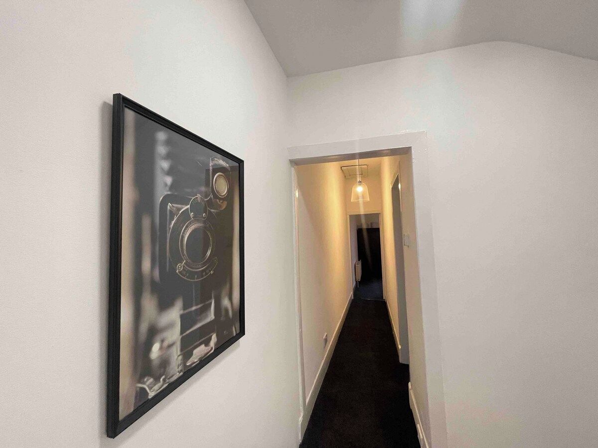 Ein Flur mit einem gerahmten Bild an der Wand, einem Teppichboden und einer Tür zu einem weiteren Zimmer.