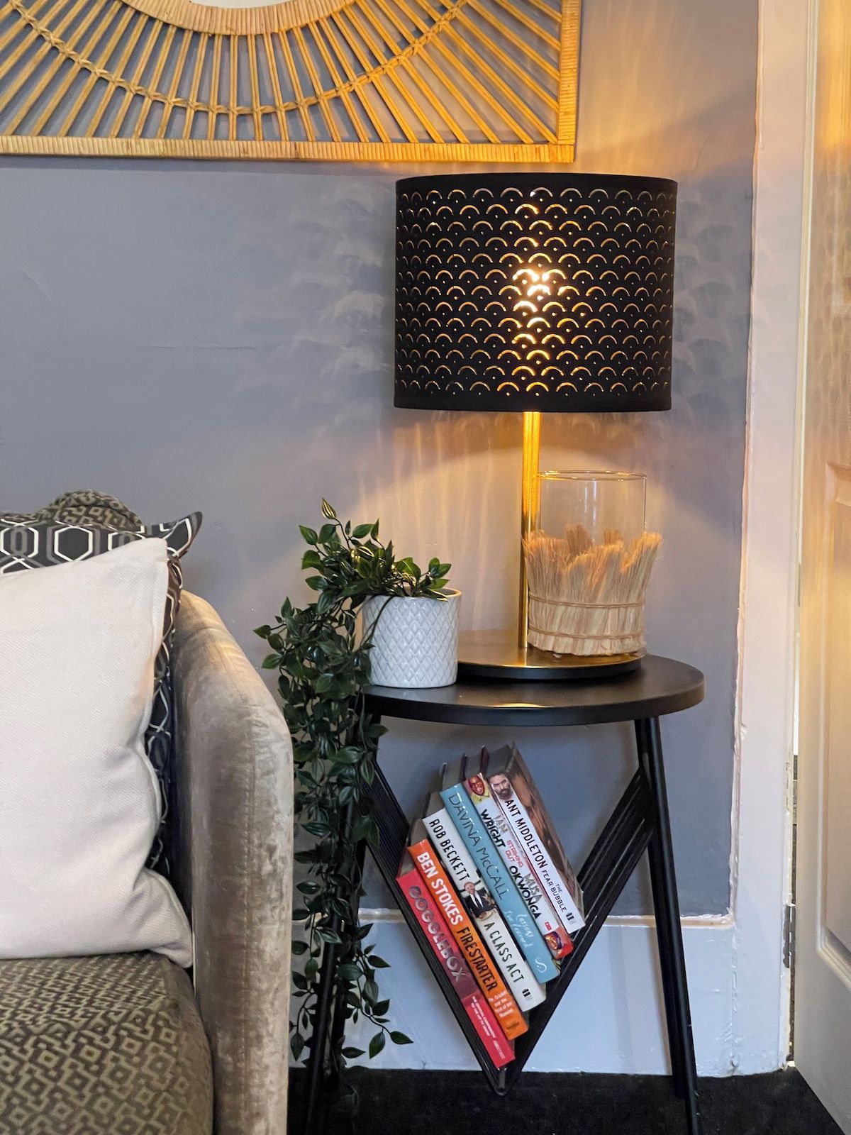 Et hjørne af et rum med en sort lampe på et lille bord, en hvid vase med grønne planter og en reol fyldt med bøger.