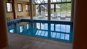 Une piscine intérieure dotée d’un sol carrelé, de murs en bois et de grandes fenêtres offrant une vue sur les bâtiments et la végétation environnants.