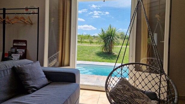 A room with a sofa, a hanging chair, and a view of a swimming pool through an open door. - Le Grand Cottage Nature Piscine Chauffée Privée (Peyrole)