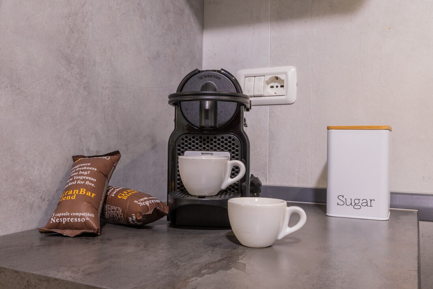 Eine Nespresso-Kaffeemaschine, eine weiße Tasse, ein Zuckerbehälter und zwei Nespresso-Kapseln auf einer Arbeitsplatte.