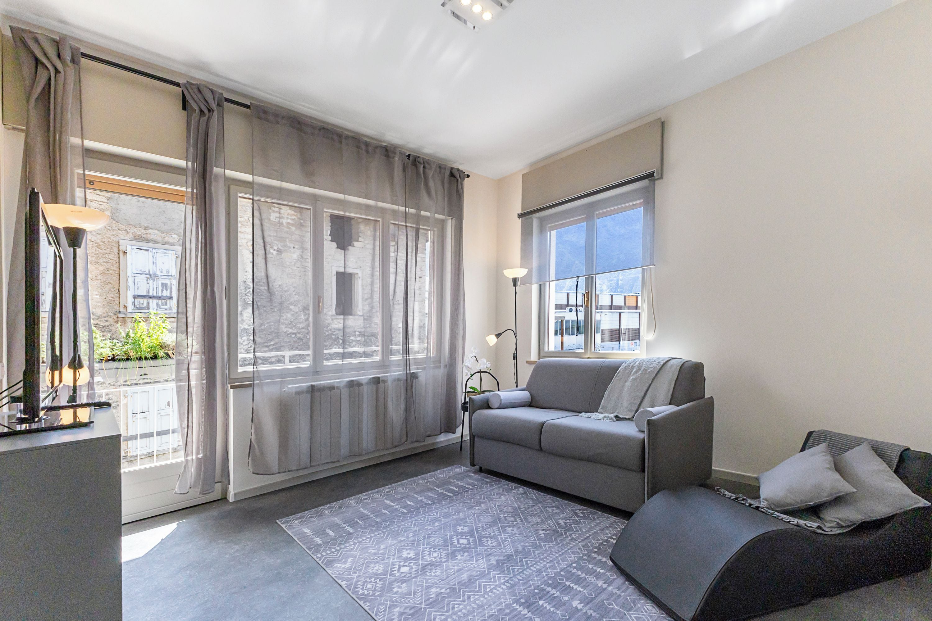A modern living room with a grey sofa, a patterned rug, and a large window with curtains.