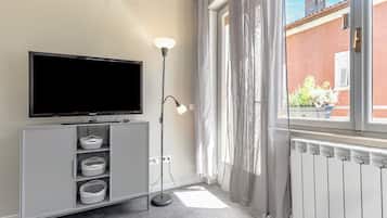 Una sala de estar moderna con televisor de pantalla plana, mueble de almacenamiento gris, lámpara de pie y vista a un edificio rojo a través de cortinas translúcidas.