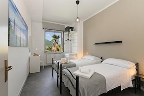 A hotel room with two beds, a small desk, a chair, a radiator, a window with a view of greenery, and a hanging light fixture.