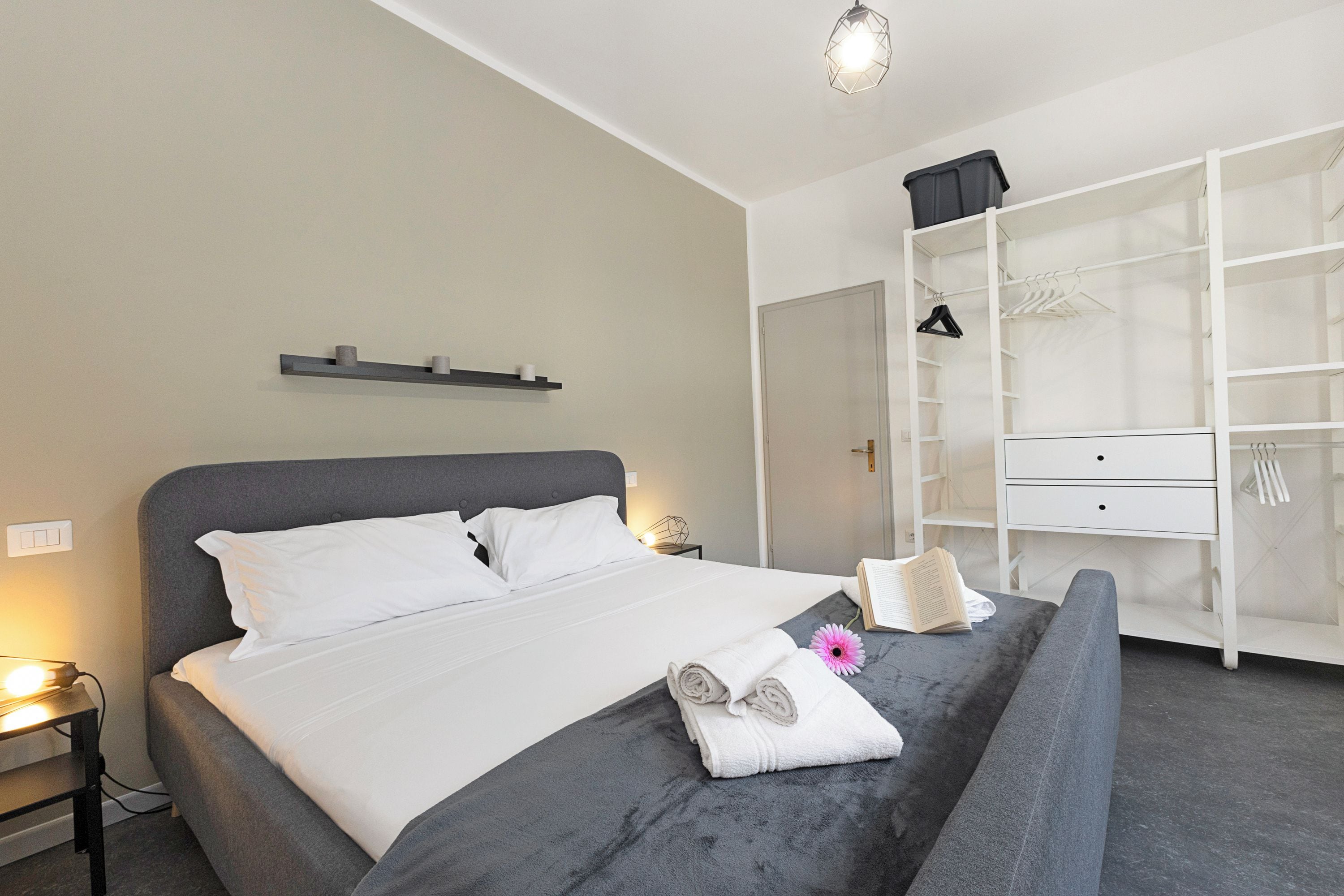 A neatly made bed with white linens, a grey headboard, and a bedside table with a lamp. A large wardrobe with multiple shelves and hanging space is visible to the right.