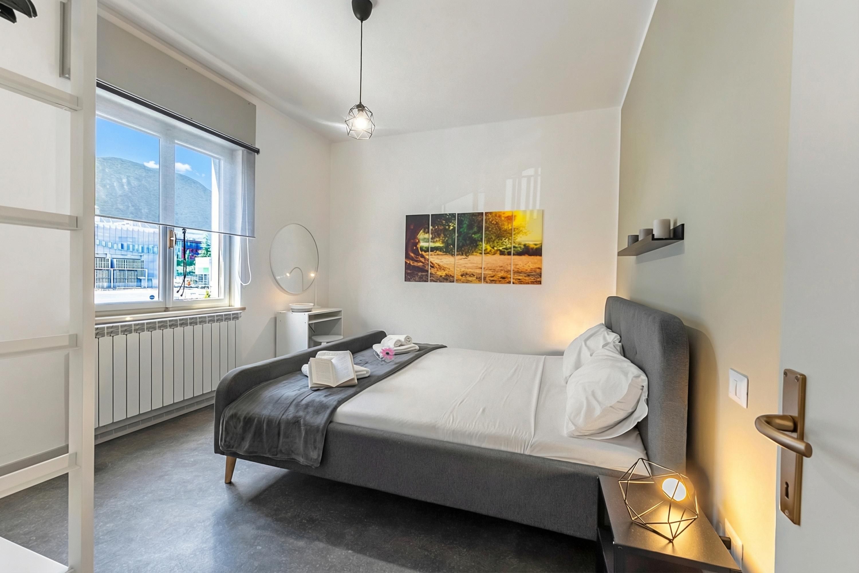 A modern hotel room with a bed, bedside table, window, radiator, and a painting on the wall.