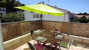 Un patio con una mesa de madera, cuatro sillas, una sombrilla verde y una pared blanca al fondo.