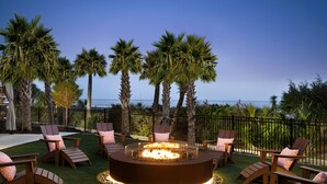 A circular fire pit surrounded by wooden chairs with cushions, set on artificial grass, with palm trees and a view of the ocean in the background. - Ocean Views in Spacious Unit at Kingston Plantation, come Relax on the Beach (Myrtle Beach)