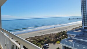 A view of a beach with a pier extending into the ocean, a parking area, and a building with a flat roof. - Ocean Views in Spacious Unit at Kingston Plantation, come Relax on the Beach (Myrtle Beach)