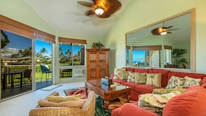 A spacious living room with a red sofa, a wicker chair, and a glass coffee table. There is a large mirror on the wall and a ceiling fan. - Grand Champions #86 Gorgeous Views On Golf Course, Near Popular Beaches (Wailea)