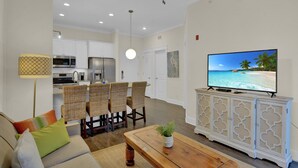 A modern living room with a sofa, coffee table, and dining area with wicker chairs. A flat-screen TV is mounted on a white cabinet, and a ceiling fan is visible. - Pet Friendly, Updated 1 Bedroom Getaway at Ocean Suites (Saint Simons Island)