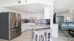 Une cuisine moderne avec des armoires blanches, un réfrigérateur en acier inoxydable et un comptoir en granit. On y trouve un tabouret de bar avec un plateau en mosaïque et un plancher de couleur claire.
